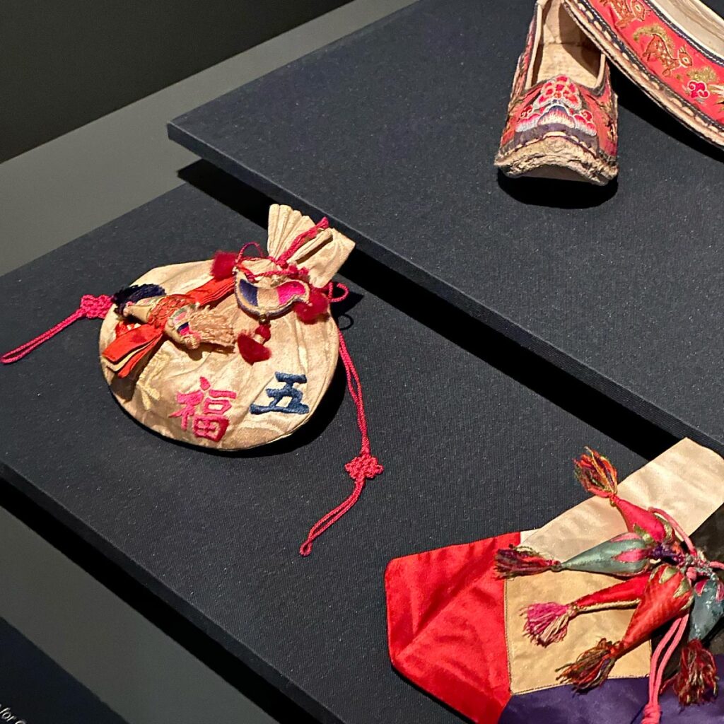 Two traditional Korean silk pouches and a single embroidered women's floral shoe displayed on a dark platform. The pouches feature silk fabric with colorful embroidery and red decorative knots, while the shoe is adorned with bright red silk and detailed needlework.