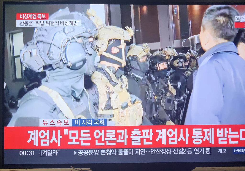 A televised news broadcast during martial law showing armed soldiers, with a caption at the bottom reading, ‘All media and publications are subject to martial law control.’