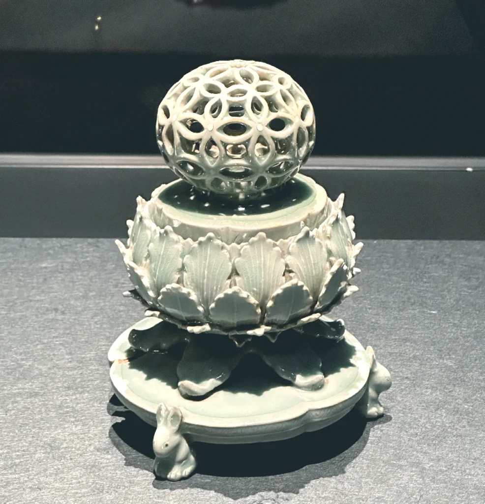 A full view of the 12th-century Goryeo Celadon Incense Burner with openwork reticulated design, a designated National Treasure of Korea. Photographed during a special exhibition at the National Museum of Korea in 2024. The artifact features an intricate, hollowed-out spherical lid patterned with interlinked circles, a body sculpted with overlapping lotus petals, and a base supported by three small, whimsical rabbits. The entire piece is covered in a mystical jade-green glaze, showcasing the pinnacle of Goryeo ceramic artistry.
