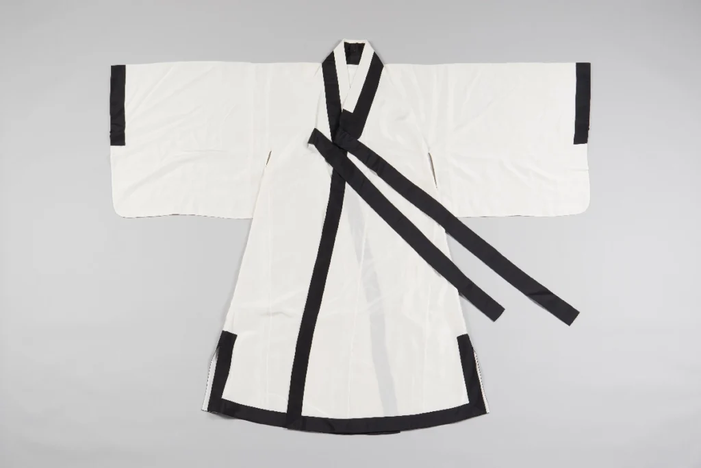 A photograph of a traditional Korean Hakchangui (Scholar's Robe). The robe is made of crisp white fabric, symbolizing the purity and integrity valued by Korean scholars. It features wide sleeves and a stark black border along the collar and edges, creating a simple yet elegant contrast. The garment is shown laid flat, representing a key example of the white-clad tradition during the Joseon Dynasty.