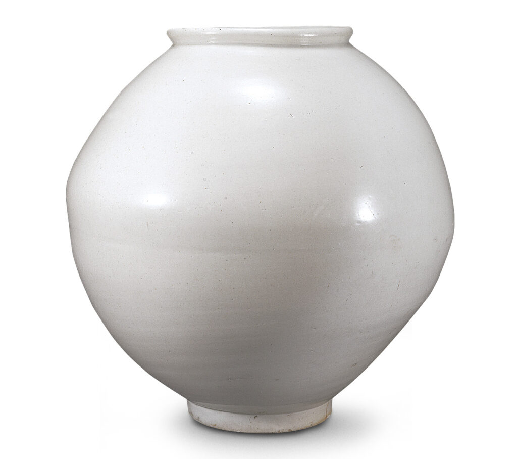 A photograph of a voluminous, spherical Korean white porcelain Moon Jar on a plain white background. The jar has a warm, creamy glaze with subtle warm stains and small firing imperfections. Its form is full and slightly asymmetrical, resting on a sturdy foot.