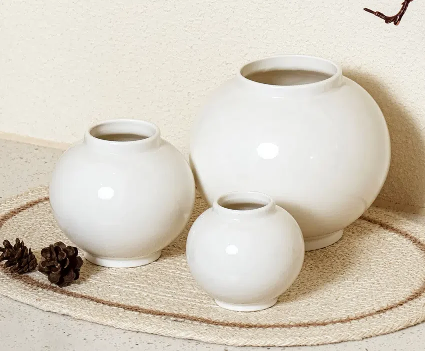 A photo of three different-sized mini Moon Jars displayed for sale in a Korean dollar store. The ivory-colored porcelain jars are arranged on a woven mat with pinecones, creating a cozy and modern interior vibe. This image illustrates how the iconic Moon Jar design has become a popular, accessible home decor item for the general public in Korea.