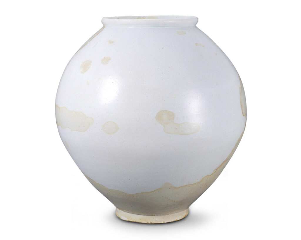 A high-resolution photograph of a National Treasure White Porcelain Moon Jar from the late 17th to early 18th century. It features a voluminous, slightly asymmetrical round body with a soft, milky white glaze. Unique yellowish-brown oil stains are naturally distributed across the surface, resulting from its historical use as an oil storage vessel. These stains create an organic, cloud-like pattern that tells the story of its practical past. (Source: National Heritage Portal)