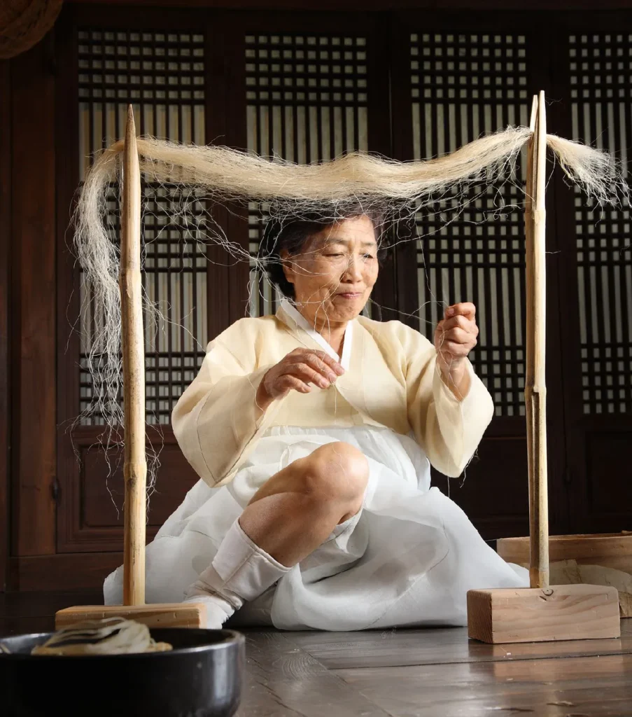 A close-up photograph capturing an elderly Intangible Cultural Heritage master artisan, dressed in a traditional silk Cream and White Hanbok and traditional White Socks, as she sits and meticulously works with fine ramie (Mosi) fibers in a traditional Korean house. Two pointed wooden stands are positioned on either side of her, from which a vast, intricate curtain of very fine, glistening ramie fibers is strung. The master has a focused expression as she holds multiple fine threads in her right hand, while her left hand is carefully guiding another single thread into the complex, veil-like web of fibers she is creating or fine-tuning. The intricate geometric patterns of traditional Korean wooden screen doors (Mun-sal) fill the background, and a dark earthenware pot with coiled ramie threads is in the foreground. The lighting is precise, emphasizing the ethereal quality of the fibers and the detailed work of the master's hands.