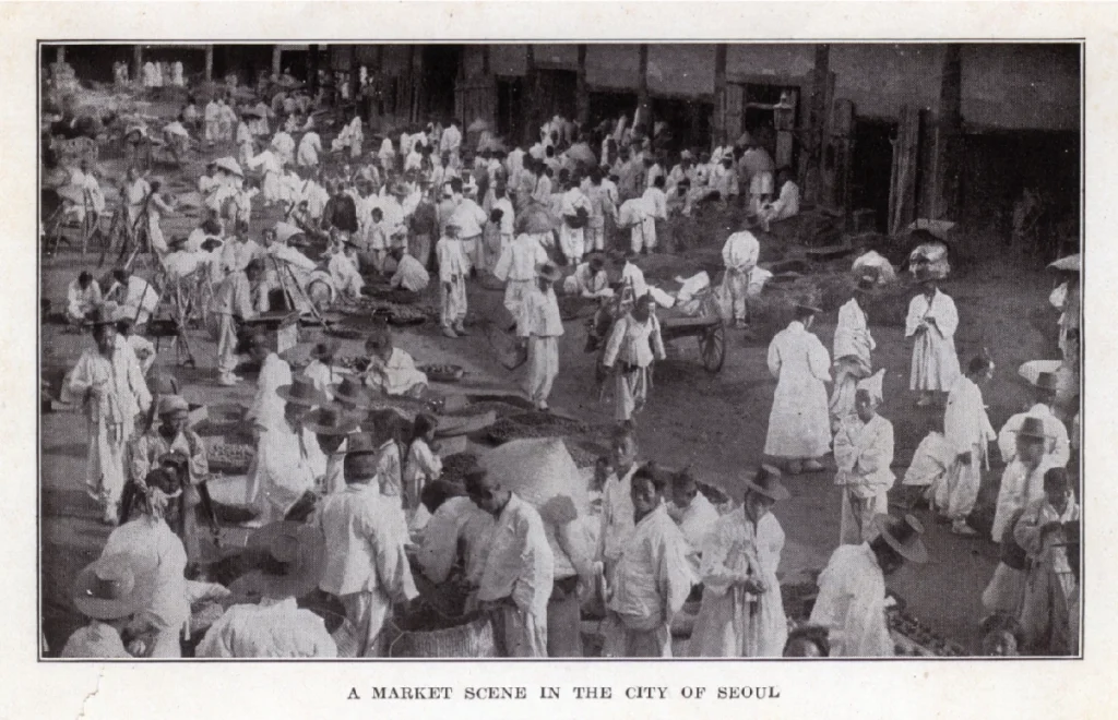 A black-and-white historical photograph titled "A MARKET SCENE IN THE CITY OF SEOUL" from around 1910. The scene captures a bustling marketplace filled with people dressed in traditional white Hanbok (Korean attire). Many men are seen wearing traditional Korean hats, such as the Gat (a black brimmed hat made of horsehair) and Paerangi (a bamboo hat). In the background, traditional tiled-roof wooden shops line the street, while merchants and customers gather around goods placed on the ground. The image reflects the lively daily atmosphere of Seoul during the early 20th century. (Source: Seoul Museum of History)