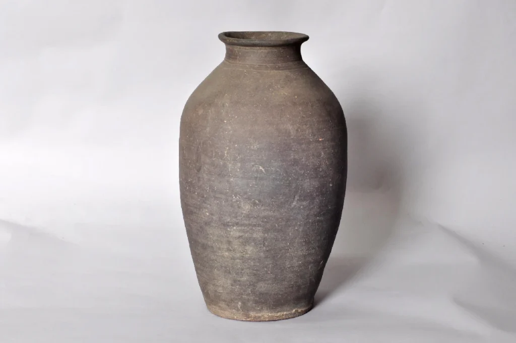 A photograph of an onggi (earthenware jar) from the Goryeo Dynasty, featuring a rich, dark greyish-brown color. The jar has a slightly out-turned rim (mouth), a wide shoulder, and a long, oval body that gradually tapers towards the base. The surface exhibits a coarse texture and subtle horizontal lines, showing the marks of time and conveying a simple, robust aesthetic.