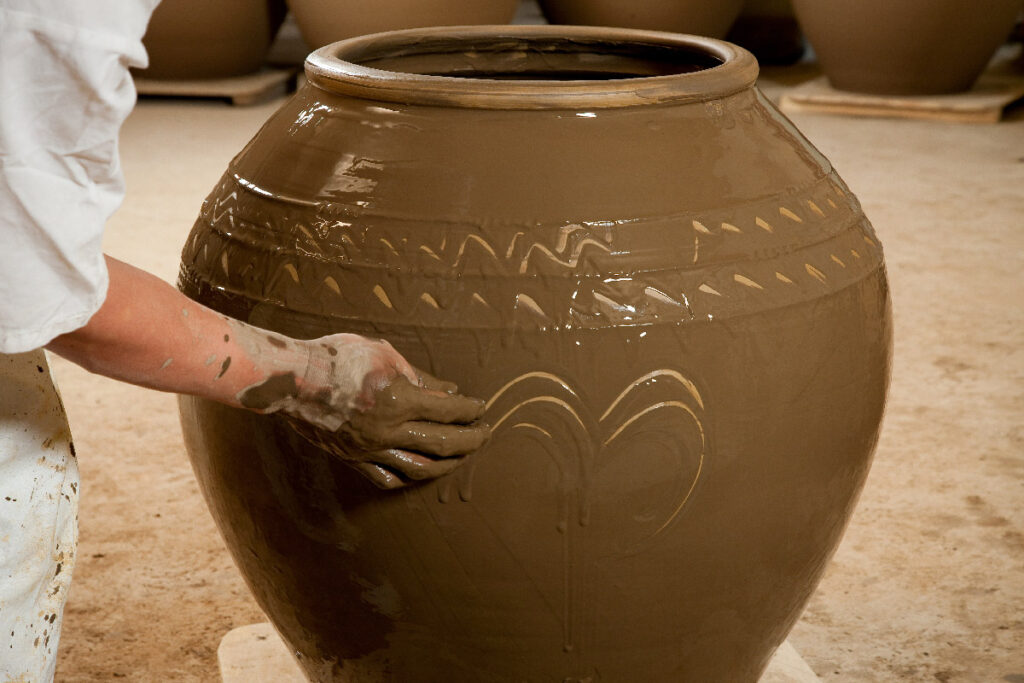 A close-up photograph capturing the hands of a skilled artisan as they perform a patterning technique on a large, wet Onggi(hangari) jar. The hand, covered in clay slips, uses finger motions to incise a delicate, arch-like pattern into the surface. The jar, made of fresh, glistening brown clay, already features other subtle, engraved wave and chevron patterns around its upper body. Multiple unfinished jars sit blurred on wooden batten supports in the background on a dusty workshop floor. The artisan’s arm in a white sleeve is visible from the left. The scene highlights the artisanal skill and tactile nature of the ceramic-making process.